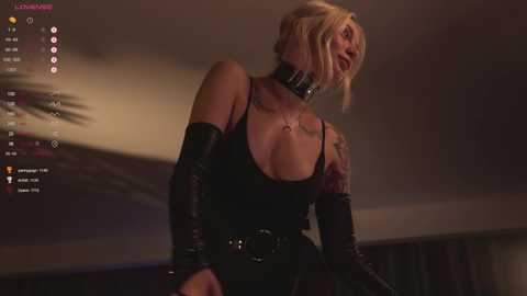 Media: Video of a blonde woman in a black latex dress and gloves, wearing a leather choker, with tattoos on her arms, set against a dimly lit background.