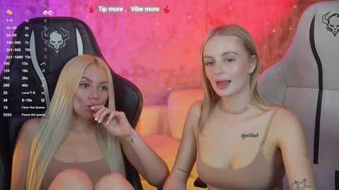 Media: A video shows two young women with light skin and long blonde hair, one wearing a tank top, the other a bralette, seated on gaming chairs. The background is a dimly lit room with pink and purple hues.