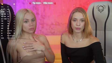 Media: Video of two young women with fair skin and long blonde hair, one topless, one wearing a black off-shoulder top, sitting side-by-side on a gaming chair in a dimly lit room.