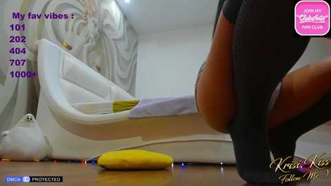 Media: A video shows a woman in a black dress, bending over a white sofa. A yellow banana lies on the floor. The room features a modern, minimalist style with grey and white decor.