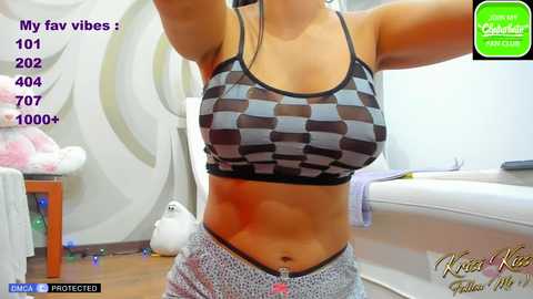 Media: Video of a fit woman with light skin, wearing a checkered sports bra and low-rise gray sweatpants, in a modern bedroom. Text overlay mentions \"My fav vibes: 101 2002 404 1000+.\