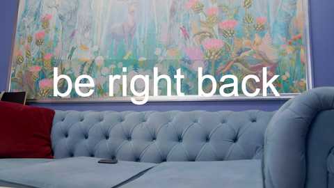 Media: A video of a light blue, tufted sofa with a red cushion against a lavender wall, featuring a large, colorful abstract painting of flowers. Text overlays the image with \"be right back.\