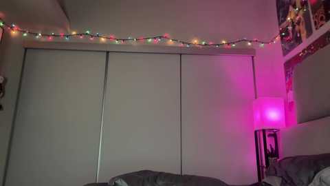 Video of a dimly lit bedroom with white sliding closet doors, string lights, a pink lamp, and a grey bed in the foreground.