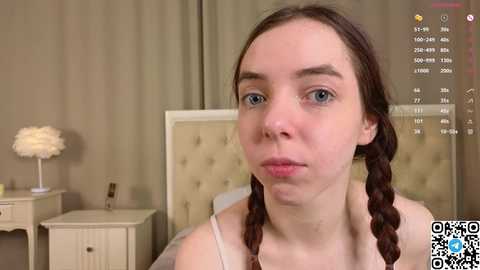 Media: Video of a young Caucasian woman with light skin and brown hair in braids, wearing a white tank top, in a beige room with a white nightstand and a cream-colored headboard.