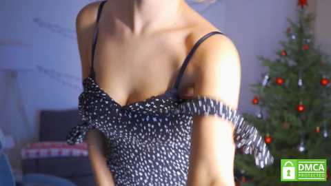 Media: A video of a slender, fair-skinned woman in a black, polka-dotted, off-shoulder dress, adjusting her straps. In the background, a Christmas tree adorned with red ornaments and twinkling lights.