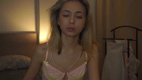 Media: Video of a young, light-skinned woman with straight blonde hair, wearing a pink and yellow lace bra, in a dimly lit bedroom with a nightstand and hanging clothes.