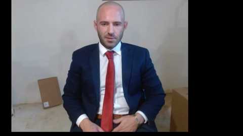 Media: Video of a bald, Caucasian man in a navy suit with a red tie, sitting in a room with cardboard boxes, wearing a neutral expression.