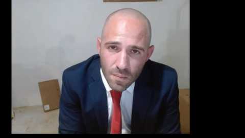 Media: A bald, middle-aged man with a short beard, wearing a dark suit and red tie, sits in a bare room with beige walls and a wooden floor, appearing contemplative.