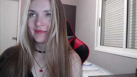 Media: A video of a fair-skinned woman with long blonde hair, wearing a black choker necklace, smiling. She sits in a gaming chair with red accents, in a room with white walls and a window with closed blinds.