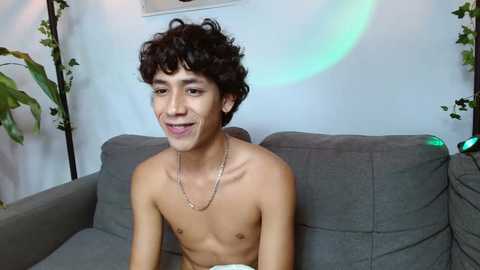 Media: Video of a shirtless, slim, young man with curly dark hair and light skin, wearing a thin gold chain necklace. He sits on a gray couch, smiling, in a modern living room with green plants and soft lighting.