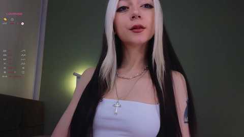 Media: Video of a pale, slim young woman with long, straight black hair and white streaks, wearing a white strapless top and gold cross necklace, indoors against a dark green wall.