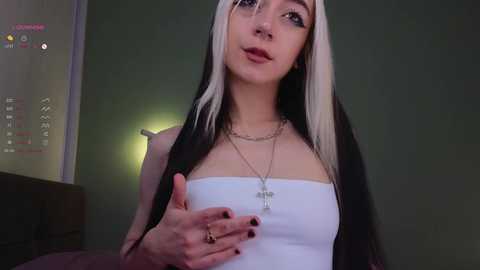 Media: Video of a pale-skinned woman with long, straight black and white hair, wearing a white strapless top and multiple necklaces, with a green wall and digital clock in the background.