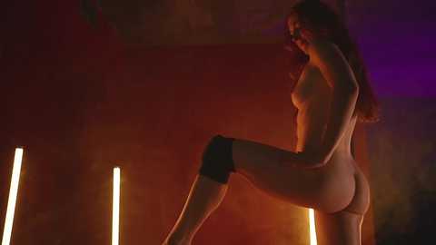 Media: Video of a nude, slender woman with long hair, wearing black knee pads, in a dimly lit room with warm, orange and purple lighting. She stands with one leg raised, creating a dynamic pose.