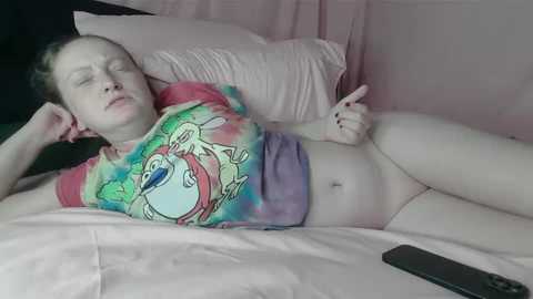 Media: Video of a pale-skinned, shirtless woman with a colorful tie-dye shirt, lying on a bed, legs apart, fingers near her crotch, with a remote control on the bed.