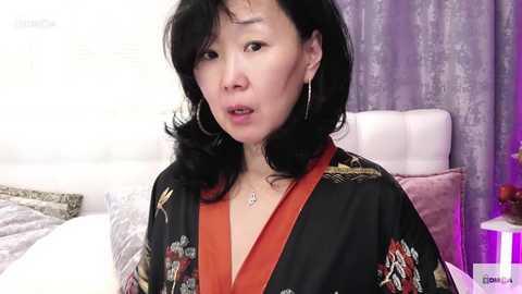 Media: Video of an Asian woman with shoulder-length black hair, wearing a red kimono with floral patterns, in a bedroom with white bedding, purple curtains, and a white headboard.