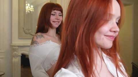 Media: Video of a young woman with long, vibrant red hair and pale skin, smiling, wearing a white lace top. She has a tattoo on her shoulder. The background features a chandelier and ornate white walls.