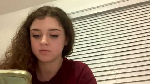 Media: Video of a young woman with curly brown hair and fair skin, wearing a maroon shirt, looking down at a green object in a room with white blinds.