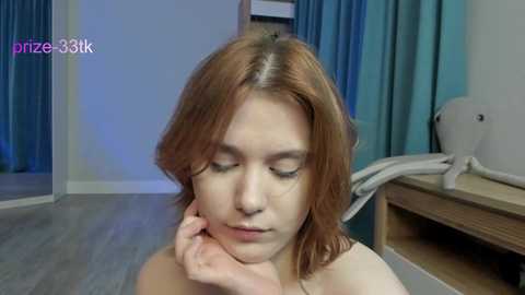 Media: A video of a young Caucasian woman with light skin and shoulder-length auburn hair, wearing a white top, resting her chin on her hand. She appears serene in a minimalist room with light wood flooring, teal curtains, and a white chair.