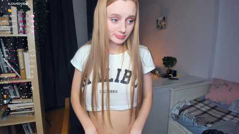 Media: Video of a slender, fair-skinned young woman with long, straight blonde hair, wearing a cropped white shirt with \"CHIBI\" printed in bold black letters, posing indoors in a cozy room with shelves and a bed in the background.