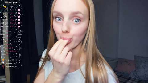 Media: Video of a young woman with fair skin, blue eyes, and long blonde hair, biting her finger while wearing a white top, indoors with a bed and digital stream overlay.