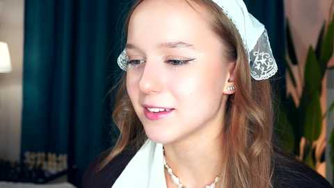 Media: Video of a young Caucasian girl with light skin, long brown hair, wearing a white lace headband, black dress, and pearl necklace, indoors with teal curtains and a green plant in the background.