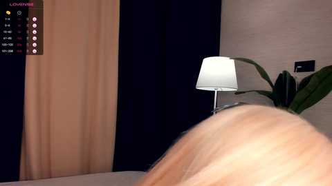 Media: A video captures a close-up of a person's bare buttocks, positioned on a bed with beige sheets, next to a modern, white lampshade. Dark curtains and a potted plant are visible in the background.