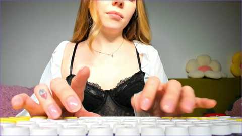 Media: Video of a young woman with fair skin, long blonde hair, and a black lace bra, typing on a white keyboard. Background includes a stuffed toy and a green shelf.