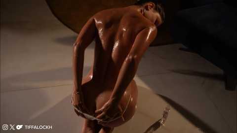 Media: A CGI image of a nude, muscular, bronzed woman with dark hair, bending over, adjusting her thong, in a dimly lit, circular room.