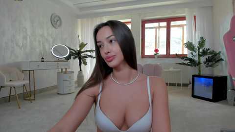 Media: Video of a slender, light-skinned woman with long dark hair, wearing a white bra, in a modern, brightly lit room with white walls, a clock, a vanity, and potted plants.