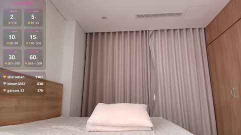 Media: A video of a minimalist, modern bedroom with beige curtains, a wooden headboard, and a neatly made bed with white sheets and a single white pillow.