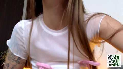 Media: A video of a young woman with long, straight brown hair, wearing a white, semi-transparent tank top that reveals her small, perky breasts and nipples. She has a tattoo on her left arm.