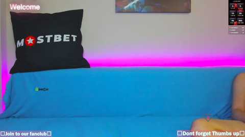 Media: A video of a modern, minimalist room with a blue couch and a black pillow with \"MISTER B\" in white and red text. A neon pink strip lights the wall behind.