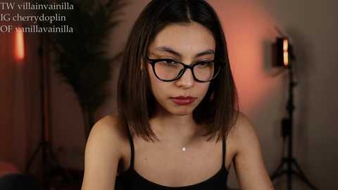 Media: Video of a young woman with straight, shoulder-length dark hair, wearing black-framed glasses and a black spaghetti-strap top. She sits in a dimly lit room with a plant and a lamp in the background.