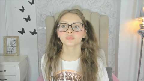 Media: Video of a young girl with long, wavy blonde hair, wearing black-rimmed glasses, a white graphic tee, and sitting in a beige chair in a whimsical room with butterfly stickers, framed artwork, and a lamp.