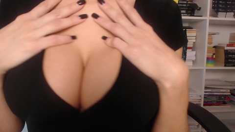 Media: Video of a woman with fair skin, large breasts, and black fingernails, wearing a low-cut black top. Background features a white bookshelf with books, binders, and a rainbow flag.