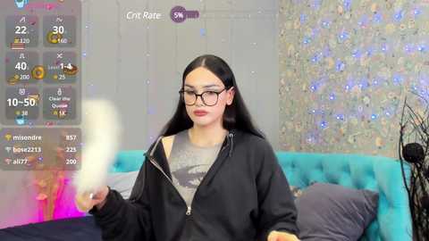 Media: A video of a young woman with long black hair and glasses, wearing a black hoodie and gray shirt, holding a white object, standing in a colorful, modern room with a blue couch and sparkling wallpaper.