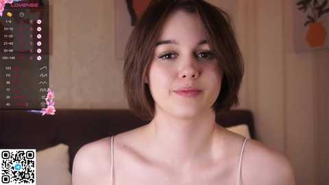 Media: Video of a young woman with light skin and short brown hair, wearing a beige spaghetti-strap top, standing in a bedroom with a dark headboard, a digital clock, and a blurred background.