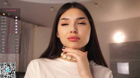 Media: Video of a young Asian woman with long black hair, closed eyes, and a slight pout, wearing a white ribbed top, and a shell necklace, indoors with blurred background.