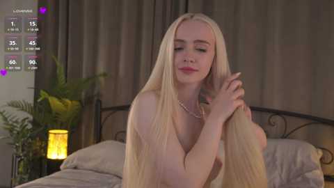 Media: Video of a fair-skinned, blonde woman with long hair, topless, sitting on a bed, touching her hair, wearing a pearl necklace, in a dimly lit bedroom.