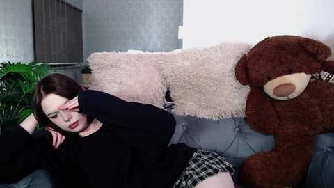 Media: Video of a young woman with fair skin and shoulder-length brown hair, wearing a black sweater and plaid skirt, resting her head on a large stuffed bear, set on a gray couch in a dimly lit room.