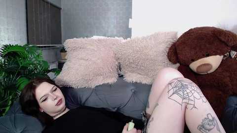 Media: Video of a young woman with pale skin, dark hair, and multiple tattoos, lying on a gray sofa, wearing a black dress. She rests her head on a fluffy pillow with a brown teddy bear beside her.