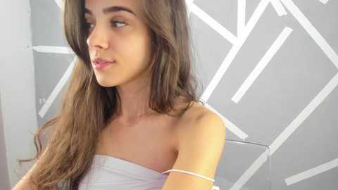 Media: Video of a young woman with long, straight brown hair, wearing a white strapless top, against a modern geometric white patterned wall. She has light skin, a slender physique, and a soft, contemplative expression.