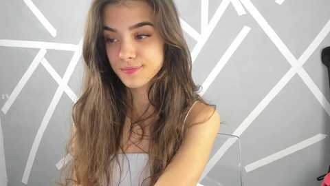 Media: Video of a young woman with long, wavy brown hair, light skin, and a slender build, wearing a white tank top. She has a soft smile and stands against a geometric white and gray wall.