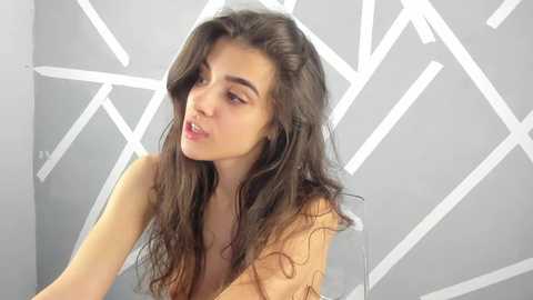 Media: Video of a young woman with long, wavy brown hair and fair skin, wearing a tan, off-the-shoulder top, against a white geometric patterned background. She has a contemplative expression.