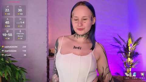 Media: Video of a fair-skinned woman with blue hair, wearing a white tank top with \"Lottie\" tattooed on her chest, standing in a room with purple lighting, surrounded by plants.