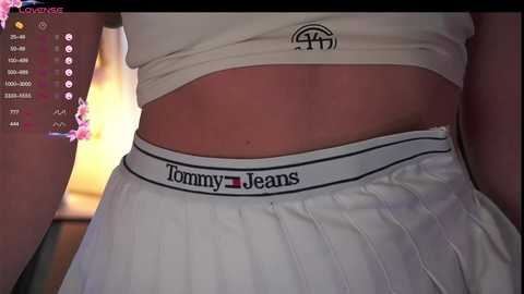 Media: Video of a woman's midsection, wearing a white Tommy Jeans ribbed tank top and matching high-waisted shorts, with the Tommy logo visible. Background features a pink floral arrangement and a video game screen with various game options.