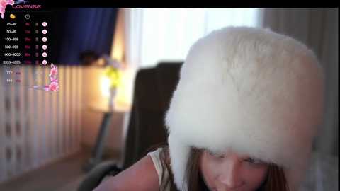 A video of a young girl in a cozy room with a fluffy white hat, wearing a striped shirt. The background shows a blurred window and a lamp.