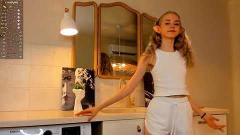 Media: Video of a young girl with blonde pigtails in a white tank top and shorts, dancing in a modern kitchen with white tiles, a mirror, and a black-and-white portrait on the countertop.