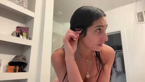 Media: Video of a young woman with long, wet black hair, adjusting it in a bathroom mirror. She wears a pink tank top and gold necklace. White shelves in background hold toiletries and a leopard print hat.