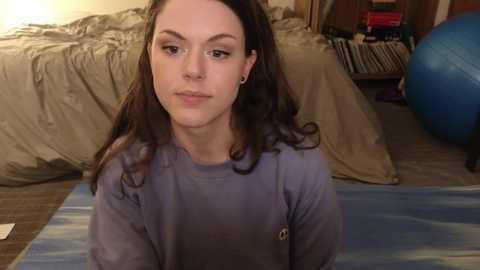 Media: Video of a young woman with long dark hair, light skin, and wearing a purple shirt, sitting on a blue yoga mat in a cluttered bedroom with a bed, exercise ball, and books in the background.